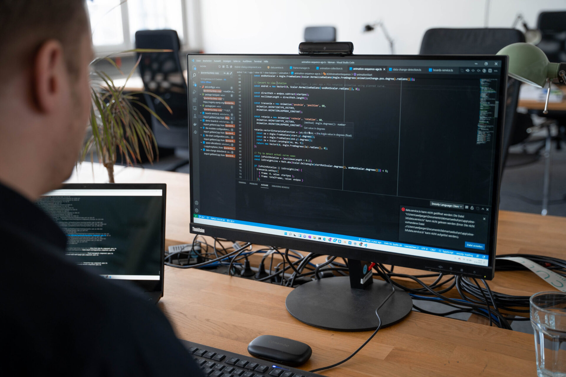 Over shoulder view of a developer sitting in front of his computer. In his screen, there is a large piece of code in a black background.