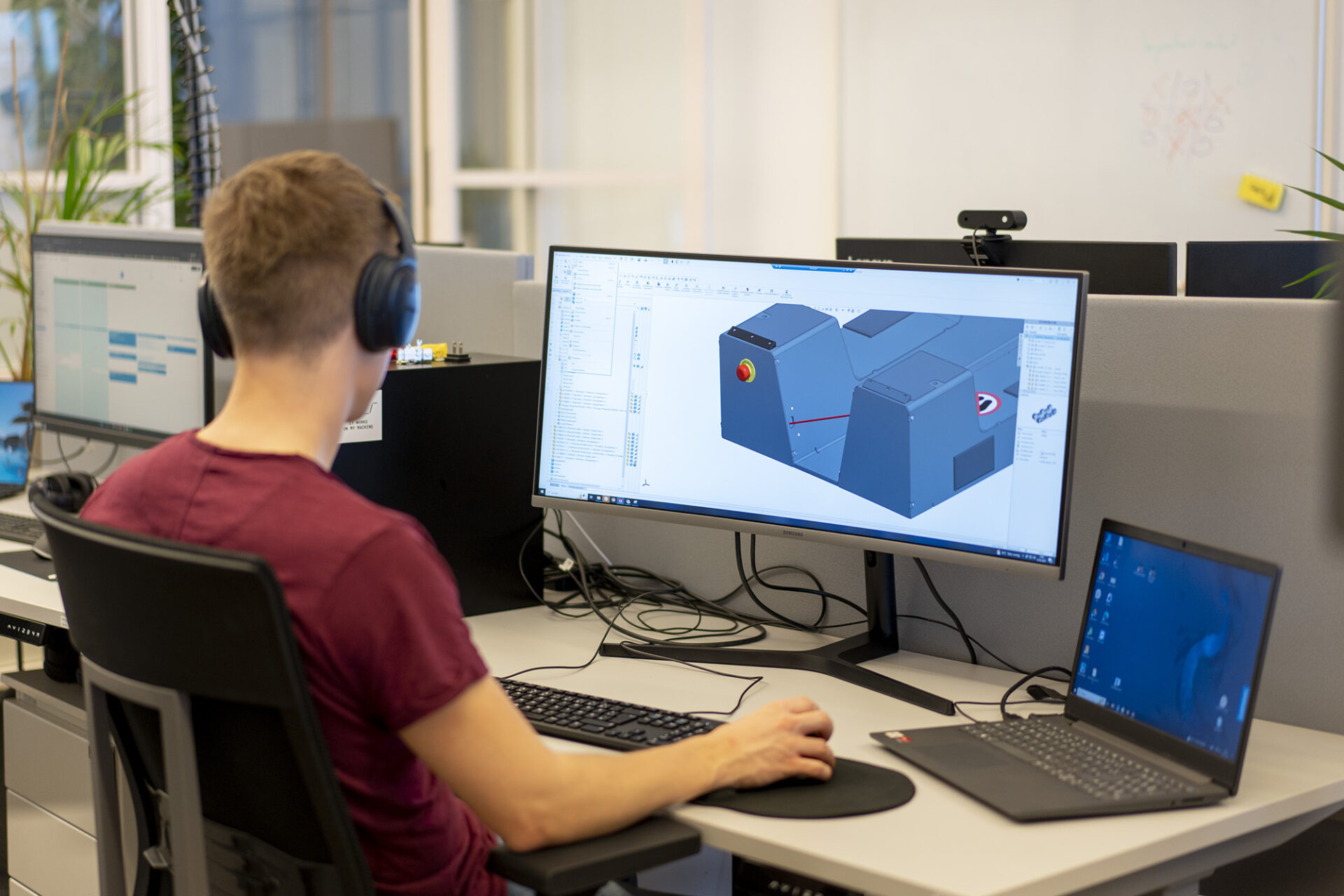 Fabian wearing a red t-shirt and sitting in front of a large monitor where a close up of a 3D rendering of a standard backpack can be seen. His back is turned to the camera. To his right is a laptop not being used, but connected to the monitor. In the background there is a white board with some calculations, which are out of focus.