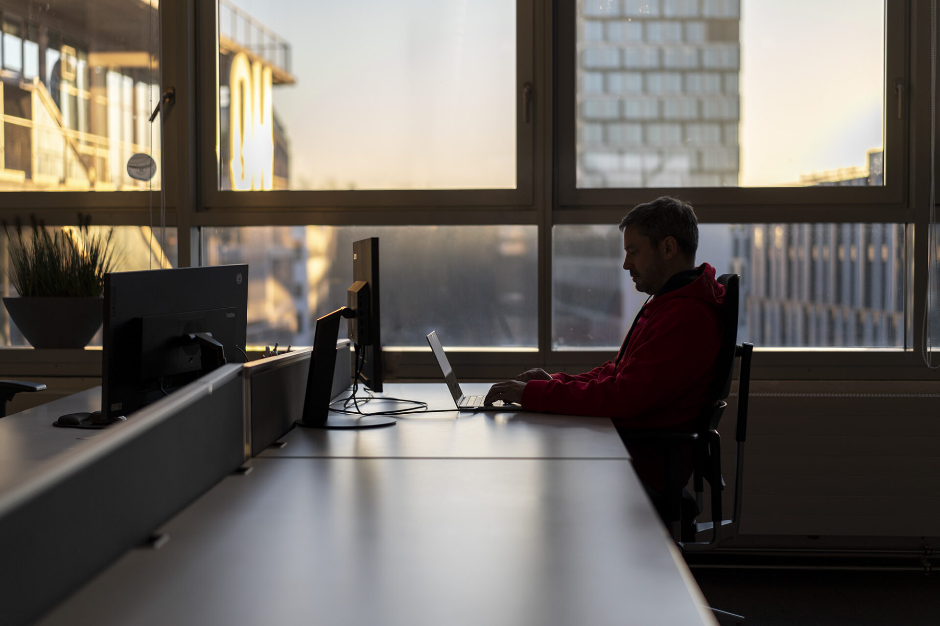 Rudolf, a dark haired men, is wearing a red jacket and sitting in front of a laptop. He has his hands on the keyboard. Behind him is a large window with sunset lighting. A few building can be seen slightly out of focus.