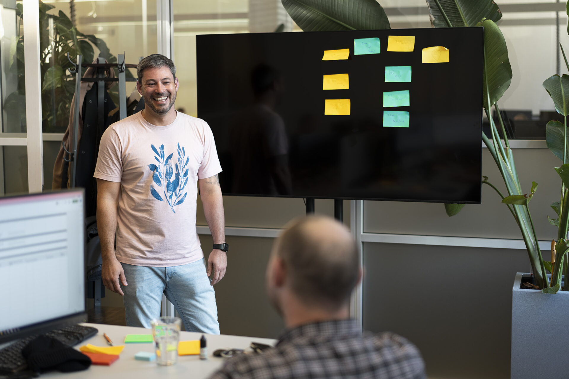 Rudolf (a dark haired men) is wearing a light pink t-shirt. Behind him and to his left is a reflective black board with 9 colourful sticky notes. In the foreground and slightly out of focus, is a balding man with his back to the camera and looking at Rudolf. He has a monitor and keyboard on his left.