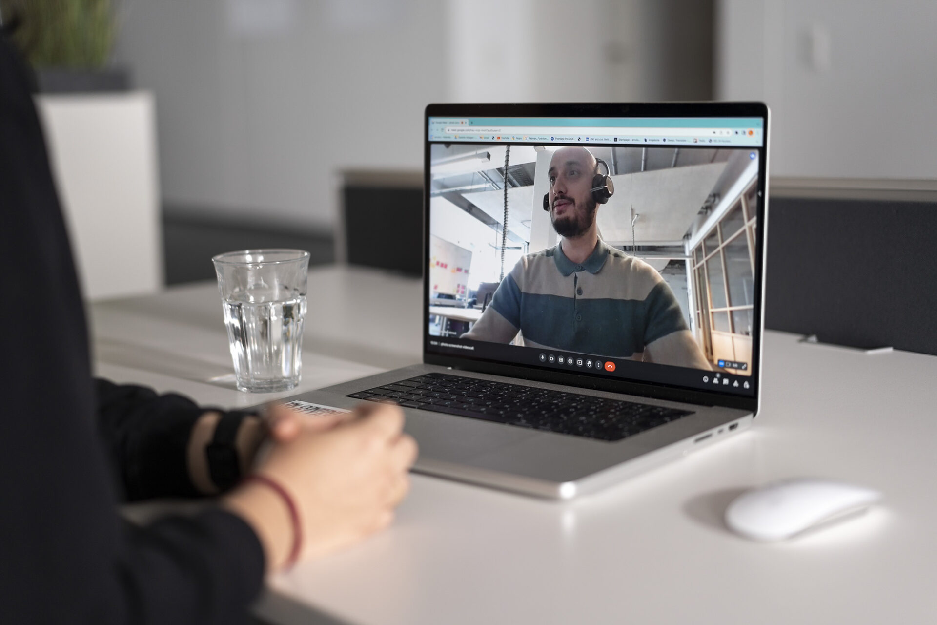 On the left, the arms of a person in a black sweater can be seen. They are crossing their hands on a white desk. In front of them is a computer. On the screen there is a videocall, where one man with headphones can be seen. 