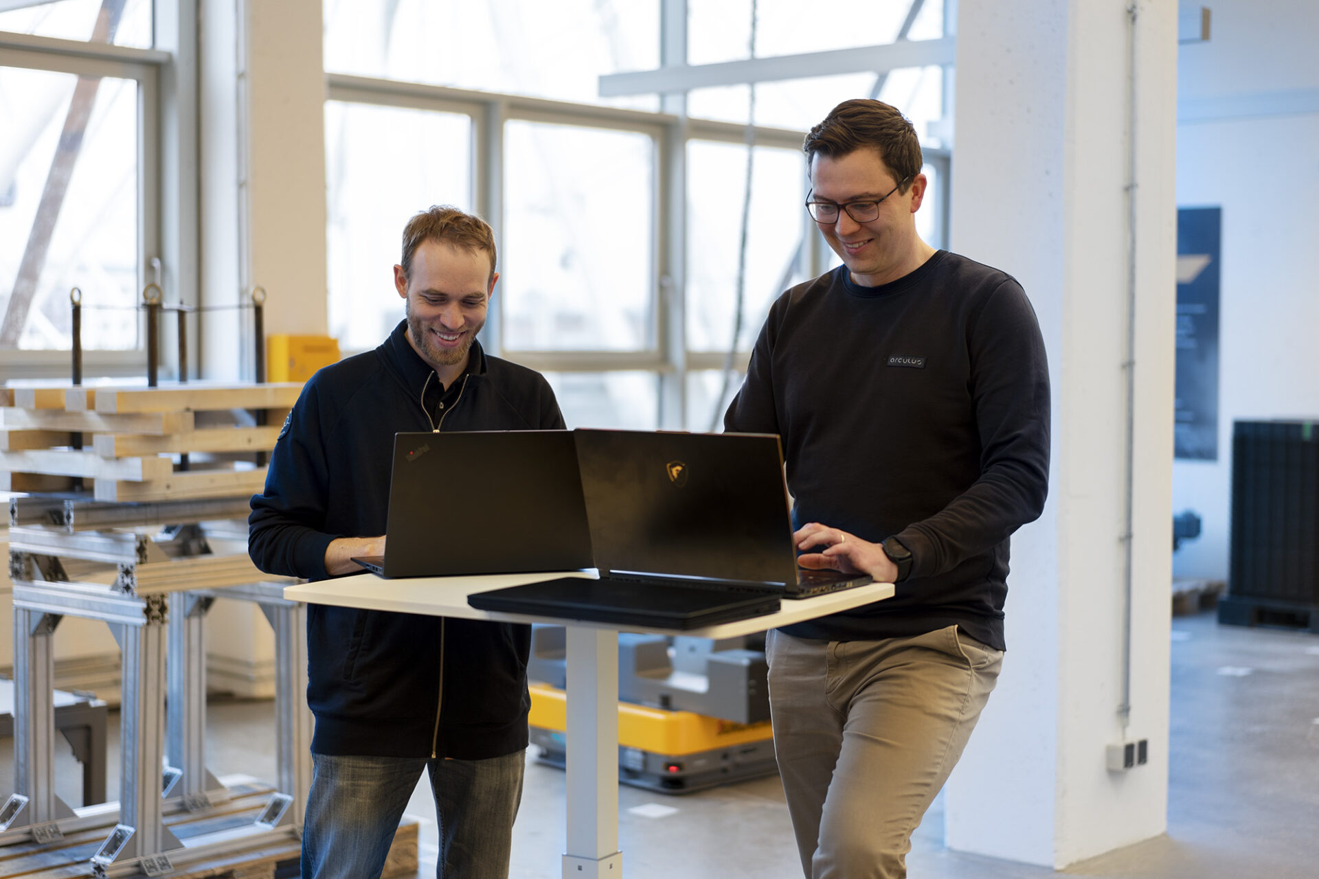 Two men are standing in a robot testing area. The man on the left is wearing a dark blue jacket. He is smiling with glasses on and has a beard. The man to the right of him is also smiling and wearing glasses. Both men are looking at their laptops.  On either side of them there are windows letting natural light into the room, illuminating their faces as they look down at their laptop screens. Behind them, on the floor, is an arculee.