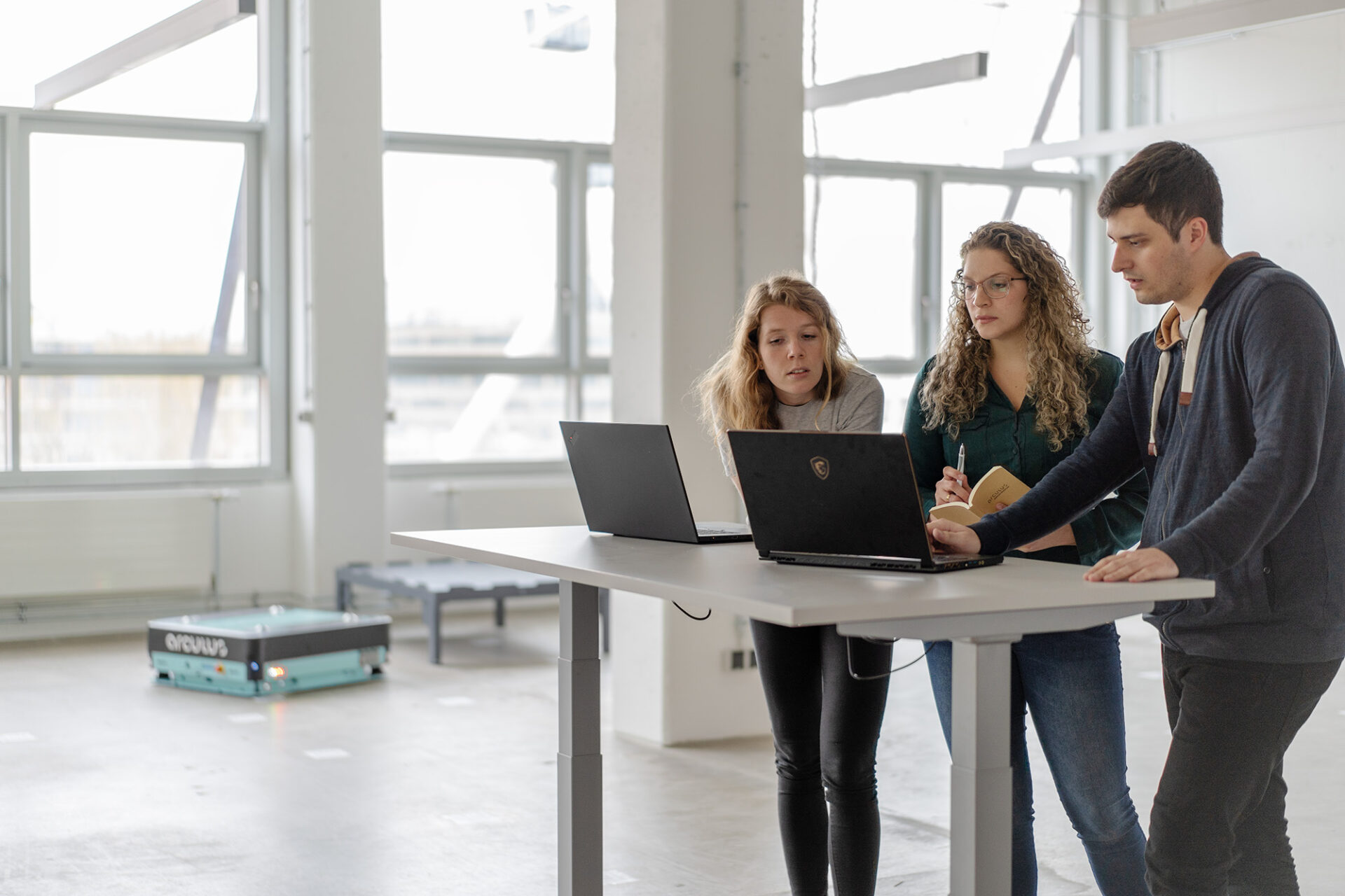 Two female engineers and one male engineer are standing in front of a high desk and looking at two laptops. In the background, there is a mint coloured arculee and a transport table.