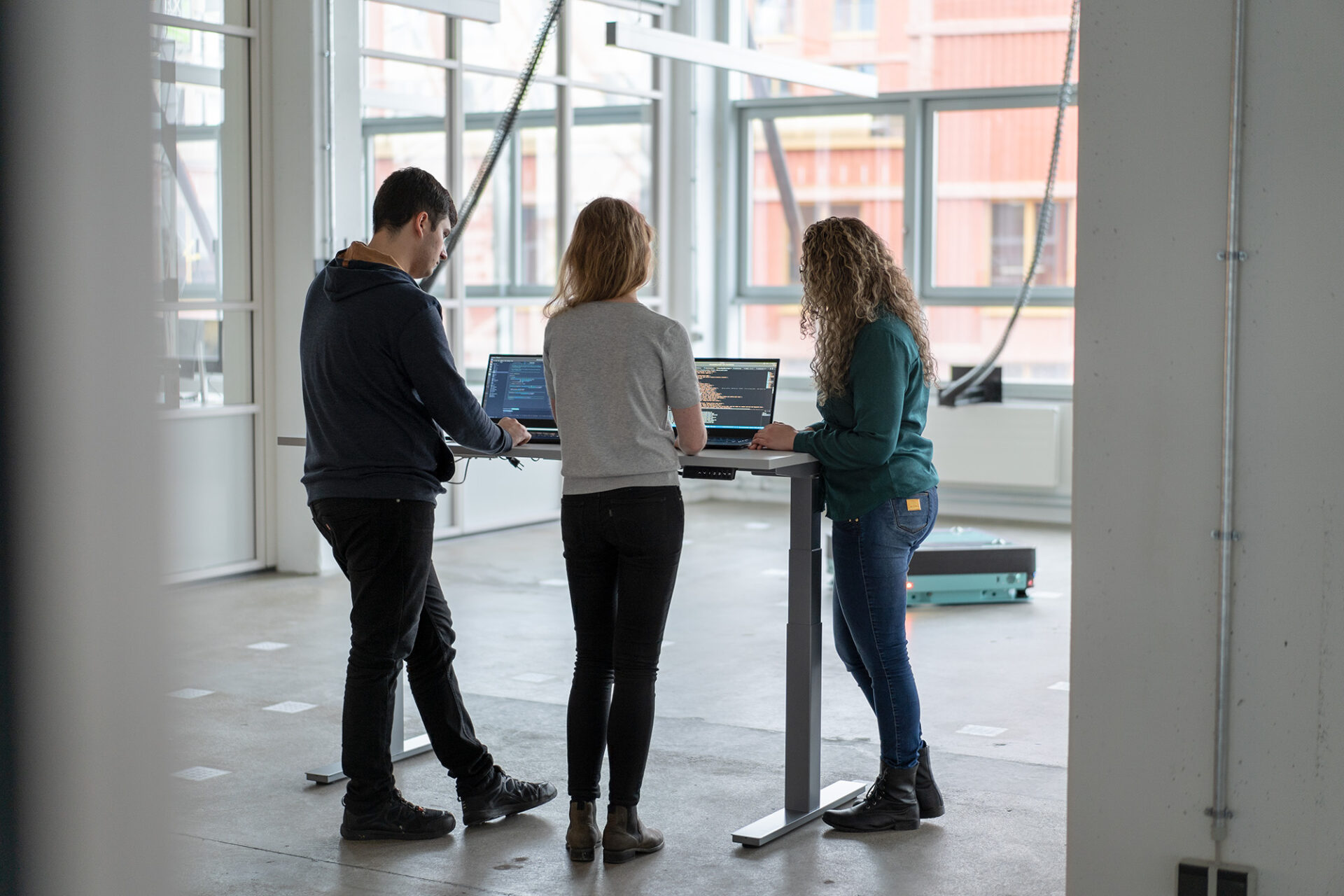 Three people standing with their backs turned to the camera. They are looking at two computer screens featuring code. From left to right: a dark haired man, a blond woman, and a brunette woman with curly hair. In the background, a green arculee is visible. 