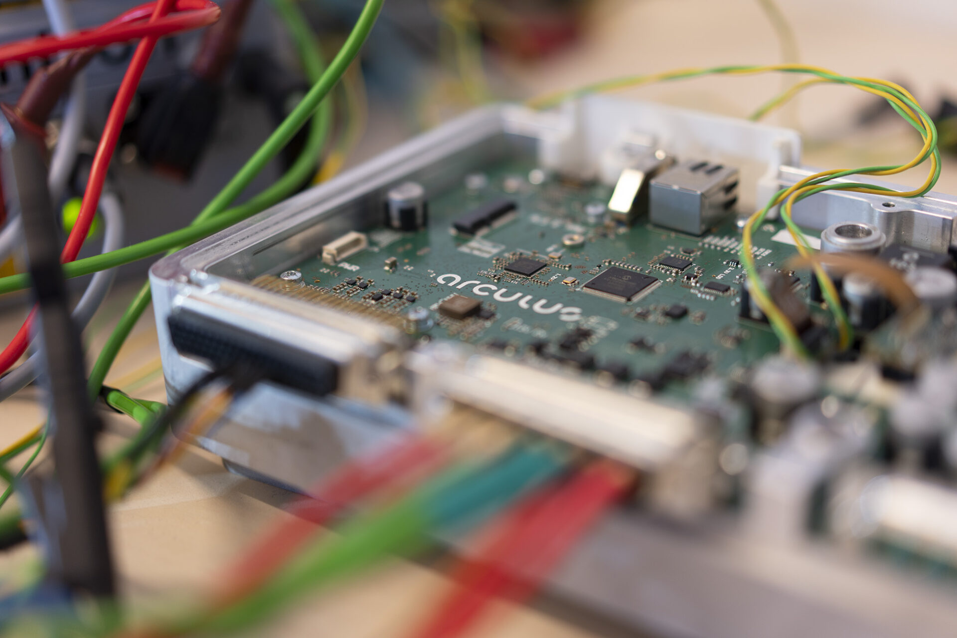 Close-up shot of electronic components and circuitry that compose the RCU® . In the center of the image, there is a circuit board with the word "arculus" displayed prominently. Surrounding the circuit board are multiple wires of different colors, including red, green, and blue wires. 