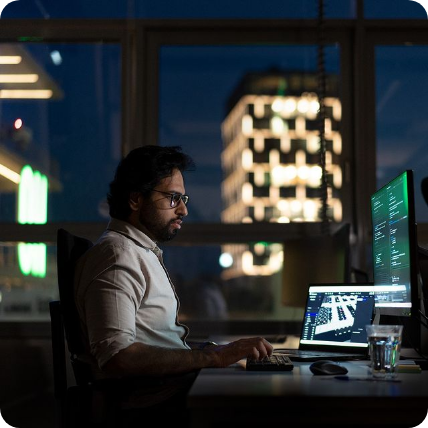 A developer working on improving our fleet manager. This is a side shot taken at night. You can see the person looking at two screen with some building in the back. Our flexible hours mean night owls are free to work whenever they feel more comfortable.