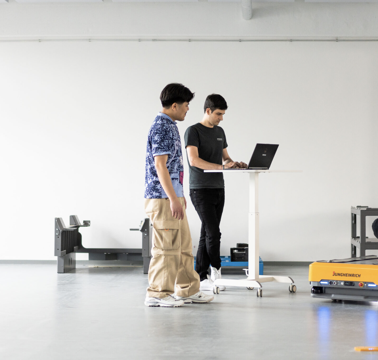 Two robotics engineers standing in front of a computer next to the arculee. Behind them is a hand-over station for pallets. The photo was taken in our in-house testing area in Munich.