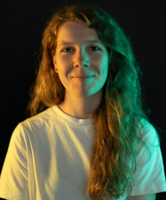 Megi is a robotics engineer at arculus. She is looking at the camera. She has a green light shining on the right side of her face and a yellow light shining on the other side.