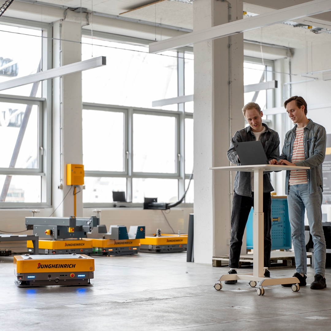 A photo of two engineers in our modern test area with some arculee Ss. They are both standing, looking at a computer while one of our modern AMRs drives close to them.